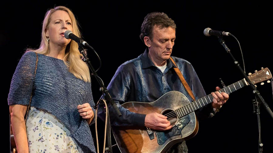 A woman sings into a microphone while a man plays guitar next to her.